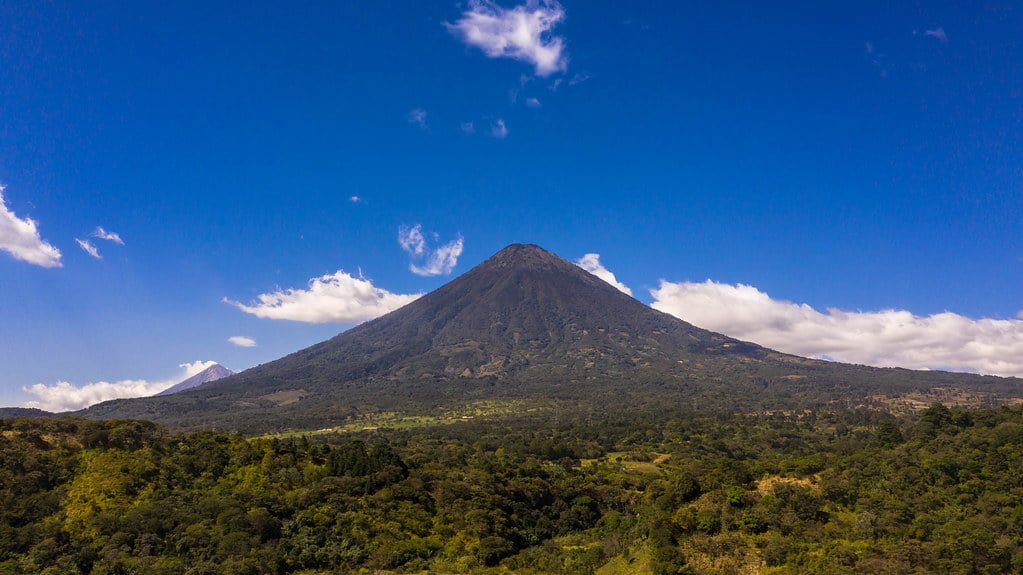 A panoramic view of a lush green landscape featuring a prominent volcano under a blue sky with sparse clouds.