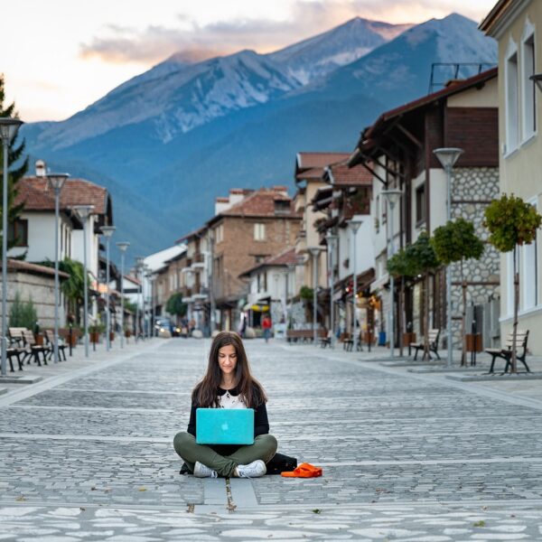 A woman sitting cross-legged on a cobblestone street using a laptop, with an empty outdoor cafe and a mountain in the background.
