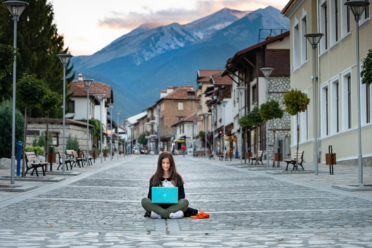 A woman sitting cross-legged on a cobblestone street using a laptop, with an empty outdoor cafe and a mountain in the background.