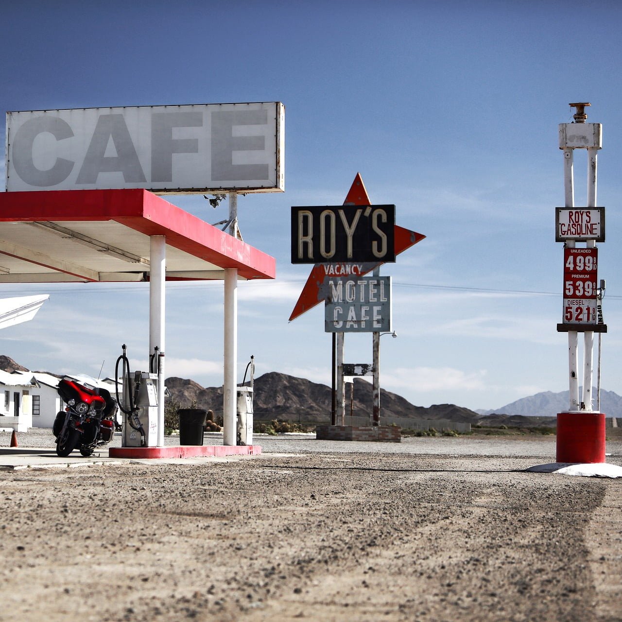 A deserted gas station with vintage signage for "CAFE" and "ROY'S MOTEL CAFE," featuring a neon star and a price board for gasoline, set against a backdrop of clear skies and distant mountains. A motorcycle is parked on the left side.
