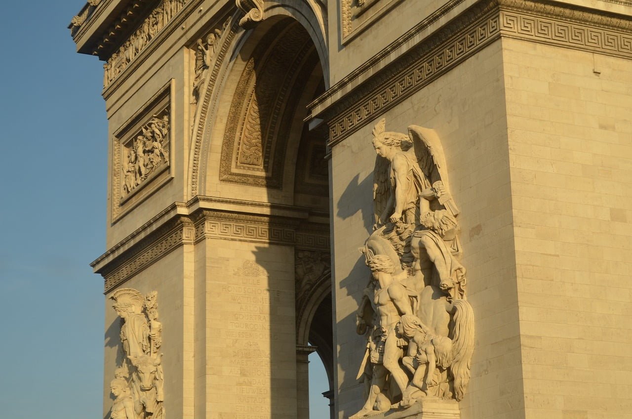 The Arc de Triomphe in Paris with intricate sculptures and engraved names of battles and cities on its facade, bathed in warm sunlight.