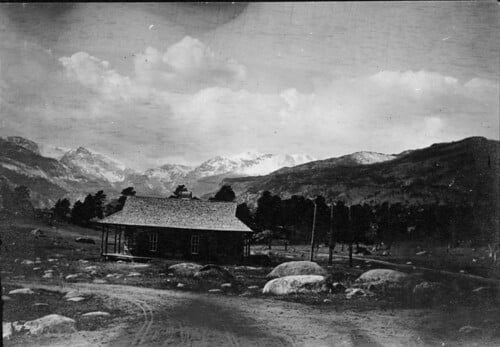 Black and white photograph of a log cabin in a mountainous landscape with scattered boulders and pine trees under a cloudy sky.