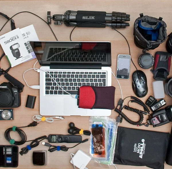 A variety of photography equipment neatly laid out on a wooden surface, including a DSLR camera, multiple lenses, a laptop, a smartphone, flash units, a tripod, cables, and other camera accessories.
