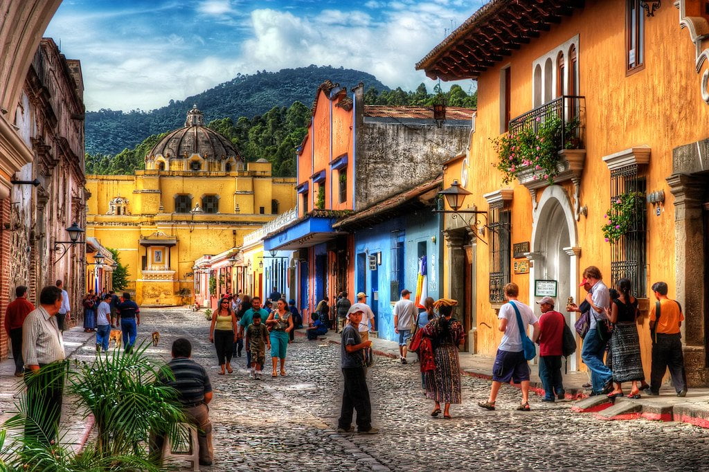 A vibrant street scene in a historic town with colonial architecture, featuring cobblestone streets, tourists and locals walking, and colorful buildings. A prominent yellow domed church is visible in the background, surrounded by lush green hills under a partly cloudy sky.