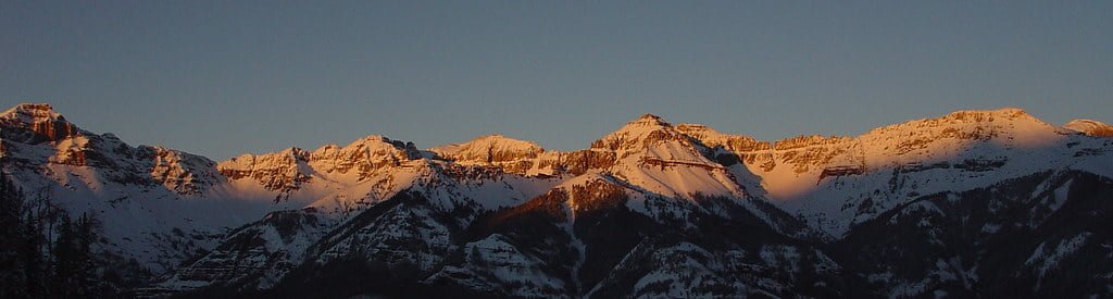 Panoramic view of snow-capped mountains at sunrise with the sunlight illuminating the peaks against a clear sky.