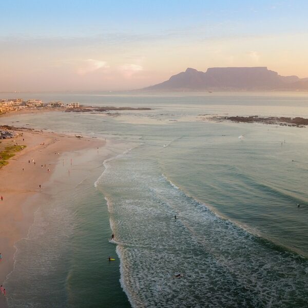 Aerial view of a beach at sunset with people walking along the shore, surfers in the water, and a view of a mountainous backdrop in the distance.
