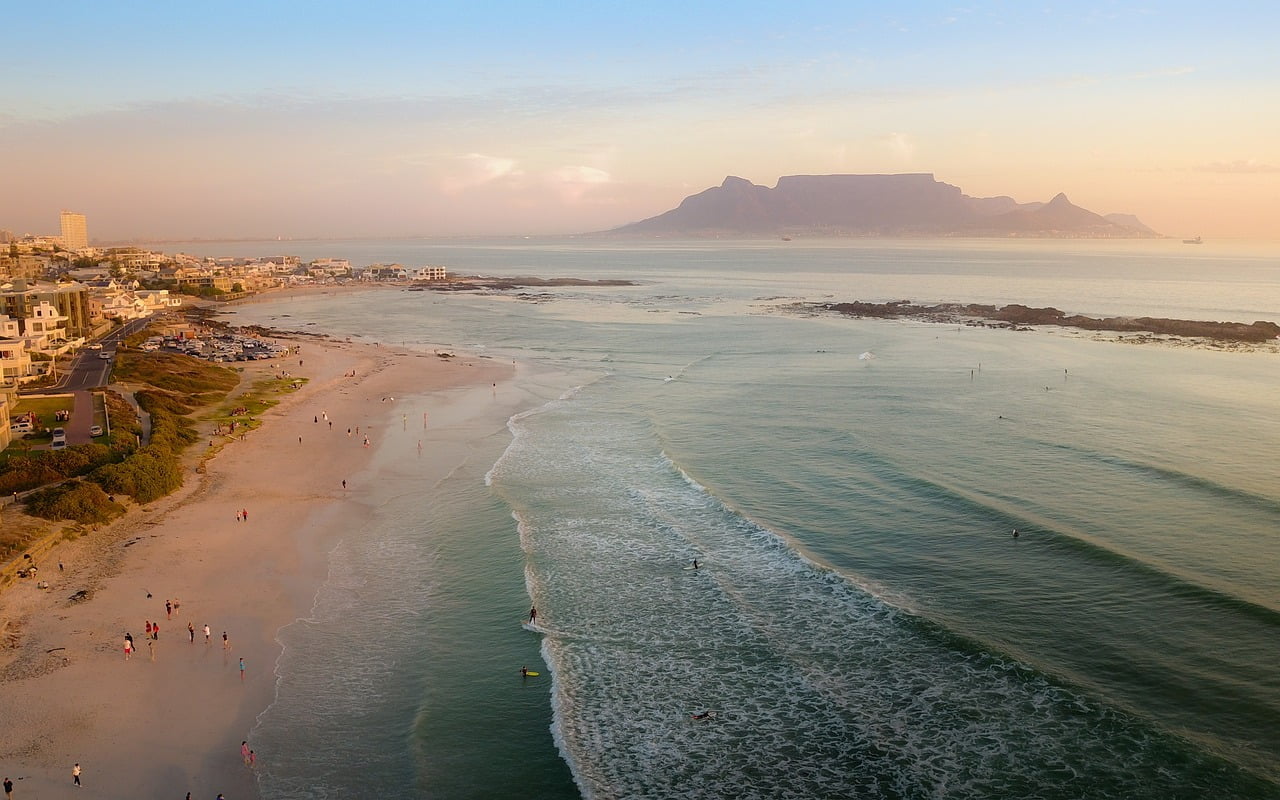 Aerial view of a beach at sunset with people walking along the shore, surfers in the water, and a view of a mountainous backdrop in the distance.