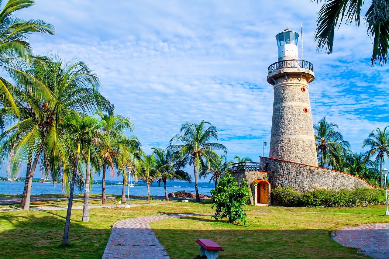 A picturesque stone lighthouse with a white lantern room and gallery, surrounded by lush green grass, palm trees, and a cobblestone path, set against a partly cloudy sky by a calm blue sea.