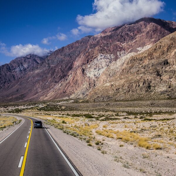 A car driving on a curving two-lane road with yellow dividing lines, surrounded by a desert landscape and rugged mountains under a blue sky partly covered with clouds.