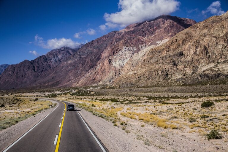 A car driving on a curving two-lane road with yellow dividing lines, surrounded by a desert landscape and rugged mountains under a blue sky partly covered with clouds.