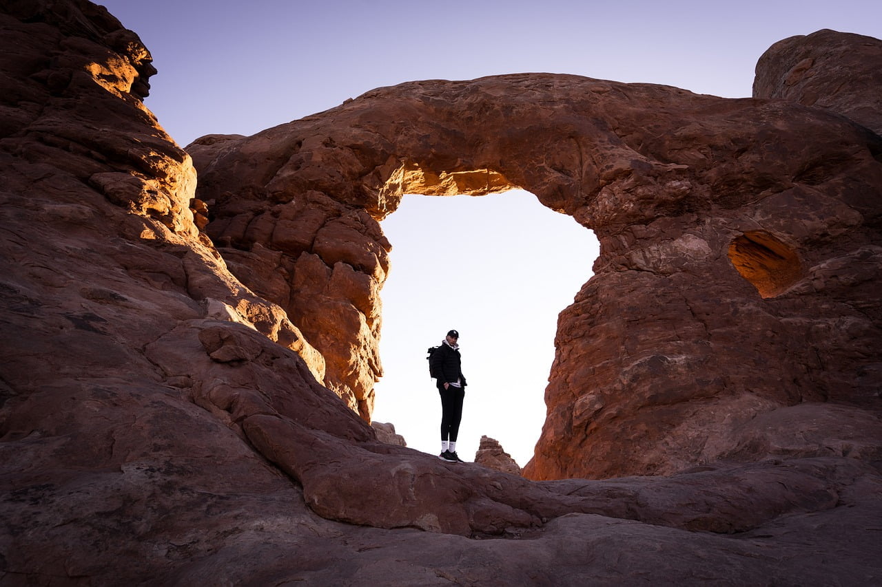 A person stands in front of a large natural rock arch, bathed in the soft light of the setting or rising sun.
