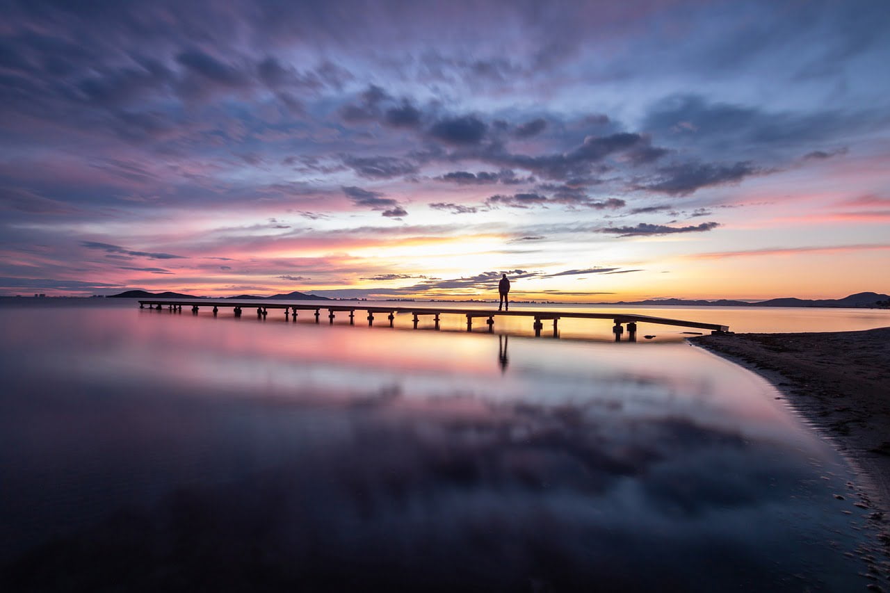 A person standing at the end of a pier during a vibrant sunset, with the colors reflecting on calm waters.