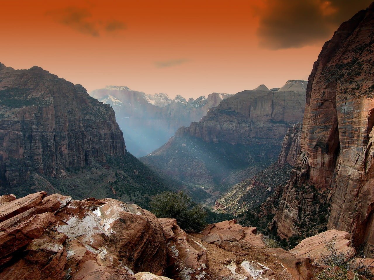 A breathtaking view of a deep canyon with layered rock formations under an orange-tinted sky, featuring sparse vegetation and patches of snow in the foreground.