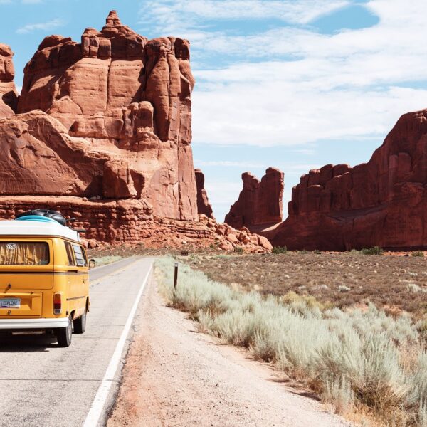 A yellow vintage van with a license plate that reads "EXPLORE" traveling on a road through a desert landscape with towering red rock formations under a clear blue sky.