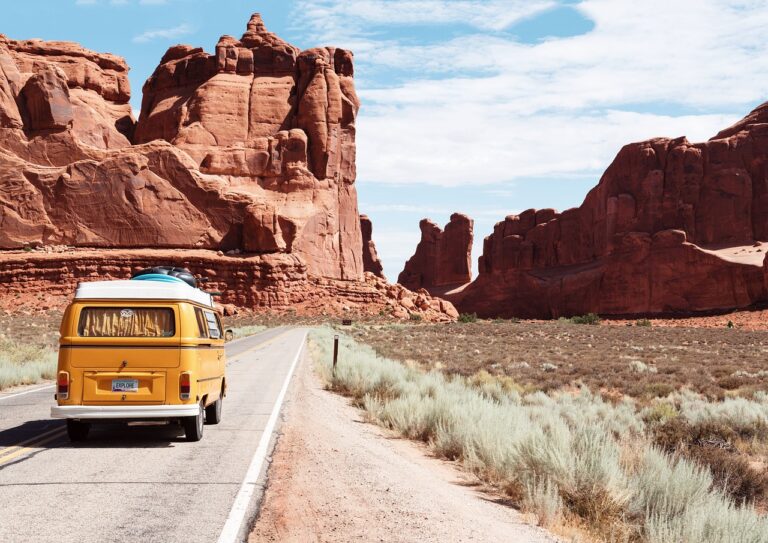 A yellow vintage van with a license plate that reads "EXPLORE" traveling on a road through a desert landscape with towering red rock formations under a clear blue sky.