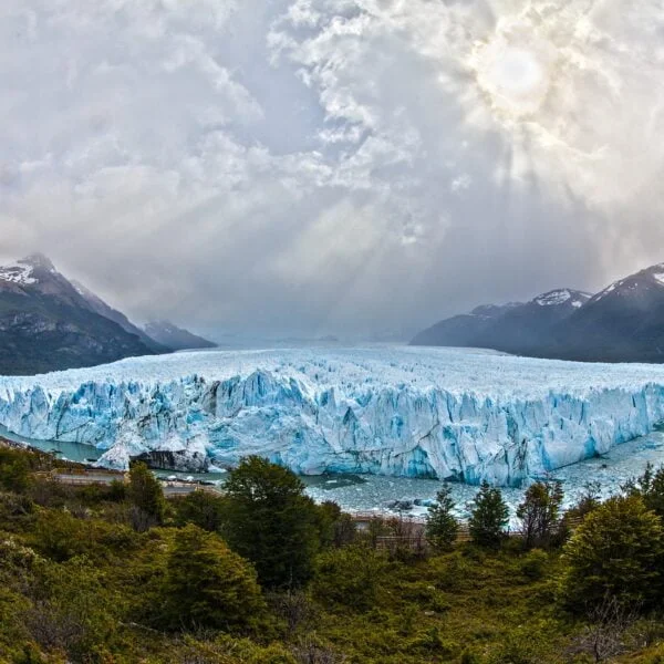 A panoramic view of the Perito Moreno Glacier with sun rays breaking through the clouds, highlighting its massive blue ice front, flanked by mountains and lush greenery in the foreground.
