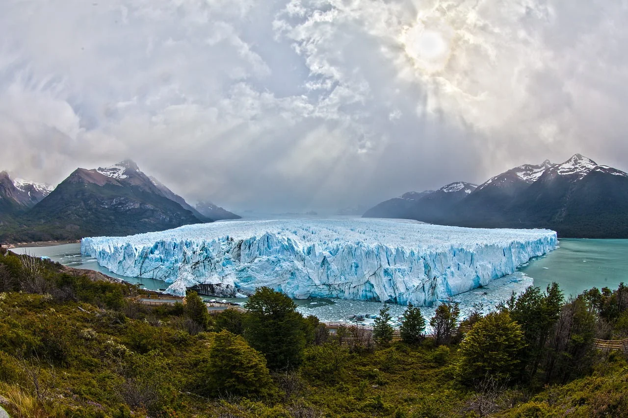 A panoramic view of the Perito Moreno Glacier with sun rays breaking through the clouds, highlighting its massive blue ice front, flanked by mountains and lush greenery in the foreground.