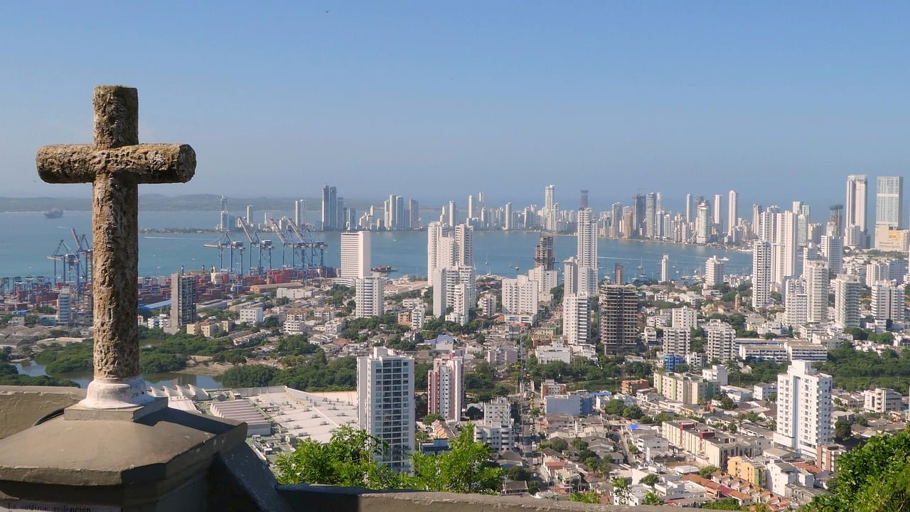 A panoramic view of a coastal city skyline from a high vantage point, featuring modern skyscrapers, a port with cranes, dense urban buildings, and a weathered stone cross in the foreground to the left.