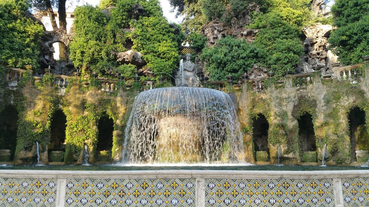 Ornate fountain with a cascading veil of water, flanked by moss-covered rocky structures and green foliage, with a tiled balustrade in the foreground.