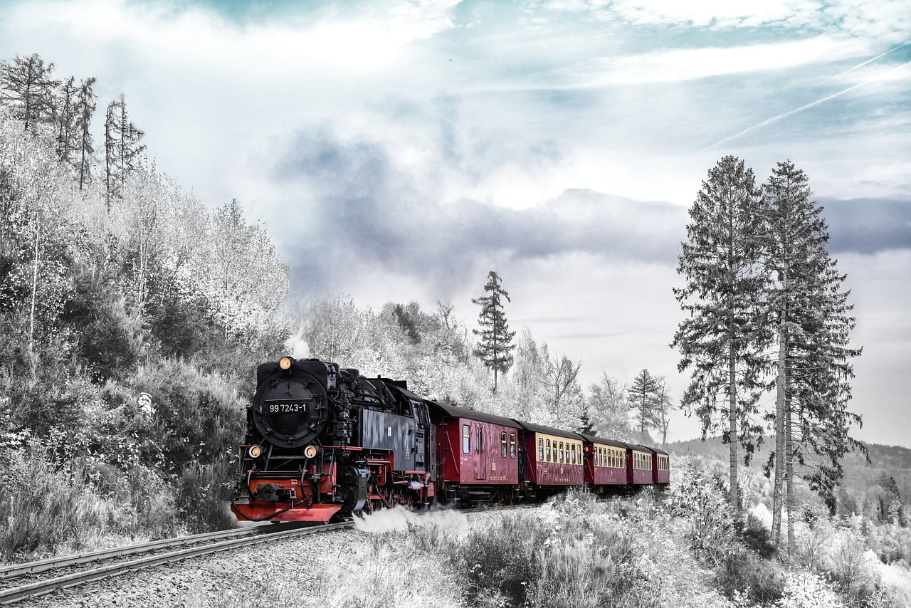 A vintage steam locomotive pulling red passenger cars through a scenic landscape with trees and cloudy skies.