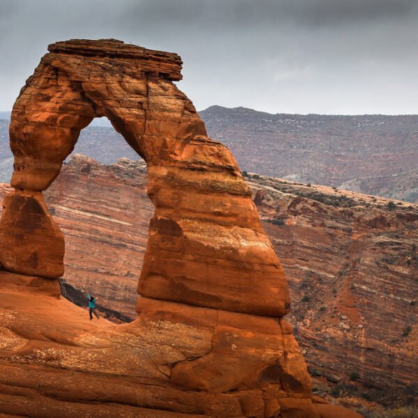 A grand natural sandstone arch with a person standing underneath it, set against a dramatic cloudy sky and hilly desert landscape.