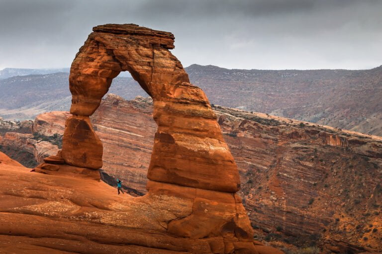 A grand natural sandstone arch with a person standing underneath it, set against a dramatic cloudy sky and hilly desert landscape.