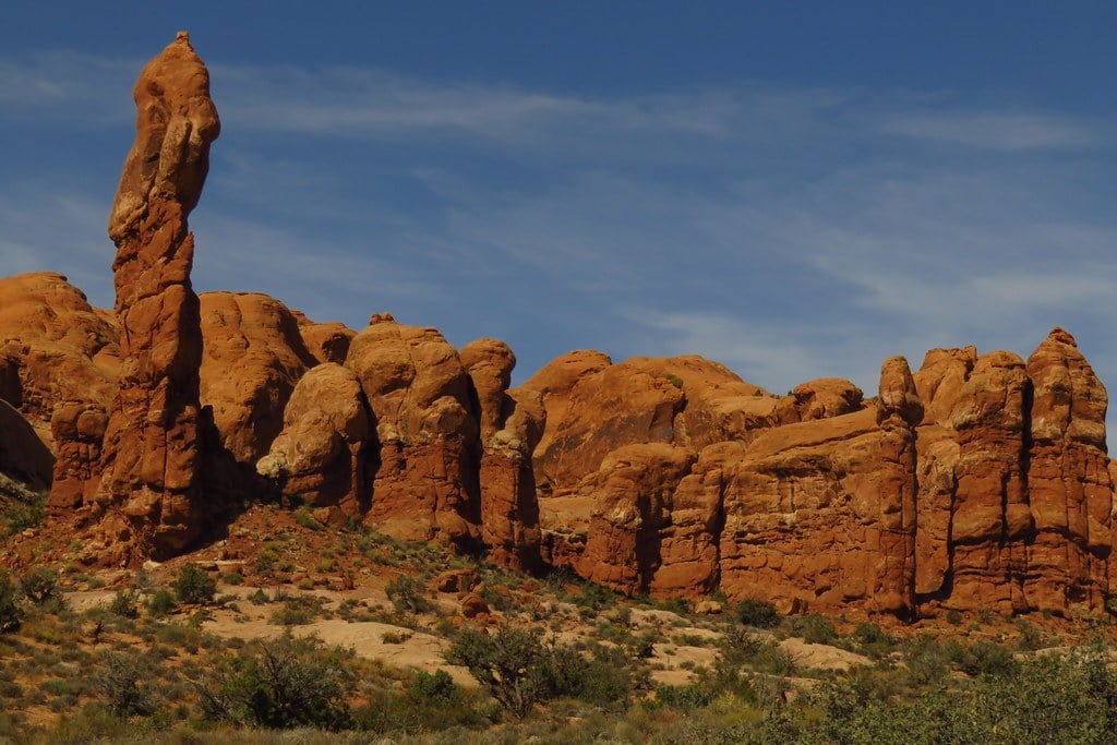 A towering red rock formation stands out amidst several large boulders under a blue sky with few clouds.