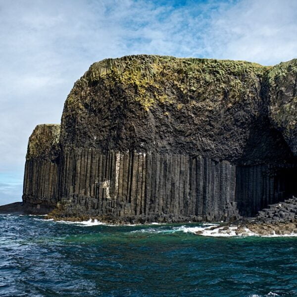 A coastal view of towering basalt column cliffs beside a body of water, with a cave-like structure visible at the base.
