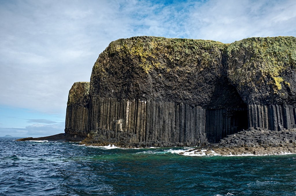 A coastal view of towering basalt column cliffs beside a body of water, with a cave-like structure visible at the base.