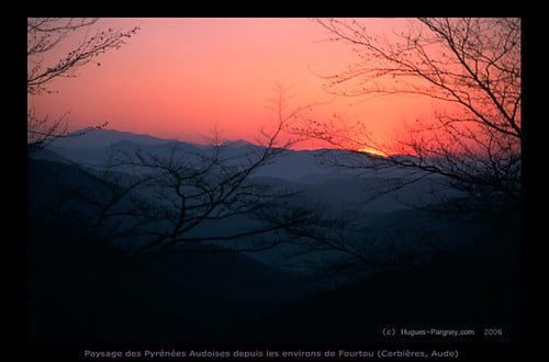 Sunset over the Pyrenees with silhouetted tree branches in the foreground and layers of mountains under a red and blue gradient sky.