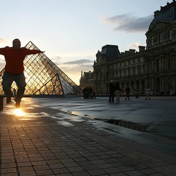 A person appears to be "leaning" against the Louvre Pyramid in a forced perspective trick photo at sunset, with the silhouette of the Louvre Museum in the background.
