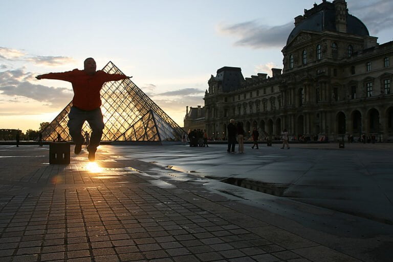 A person appears to be "leaning" against the Louvre Pyramid in a forced perspective trick photo at sunset, with the silhouette of the Louvre Museum in the background.
