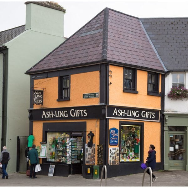 A street view of a quaint orange building labeled "ASH-LING GIFTS" next to a green building with a "BLOUSERS" shop, with pedestrians walking by and hanging flower baskets adorning a lamppost.