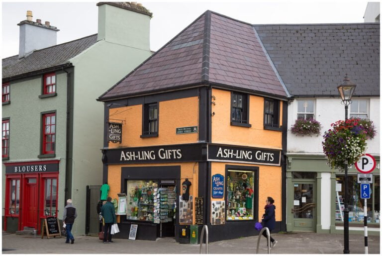 A street view of a quaint orange building labeled "ASH-LING GIFTS" next to a green building with a "BLOUSERS" shop, with pedestrians walking by and hanging flower baskets adorning a lamppost.