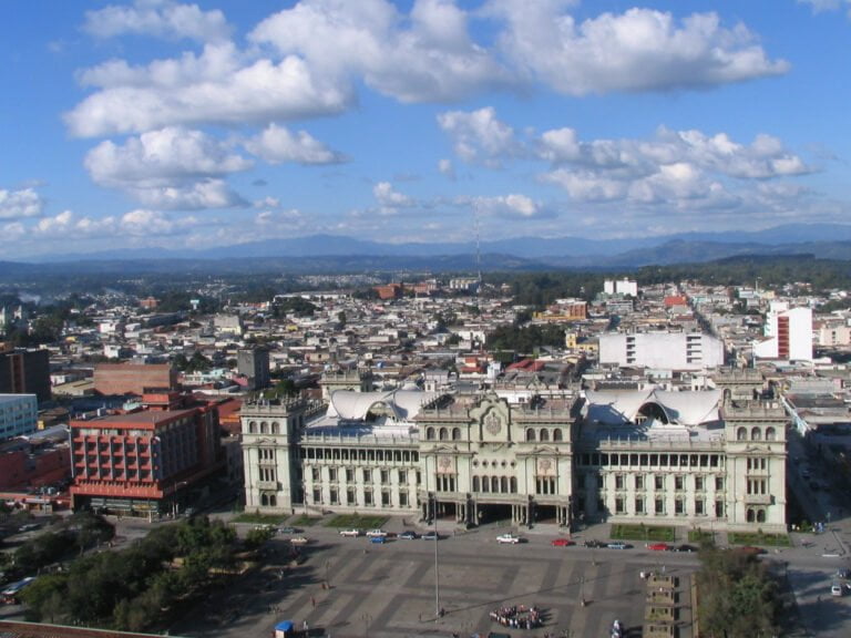 A panoramic view of a cityscape with an ornate building in the foreground and scattered clouds above.