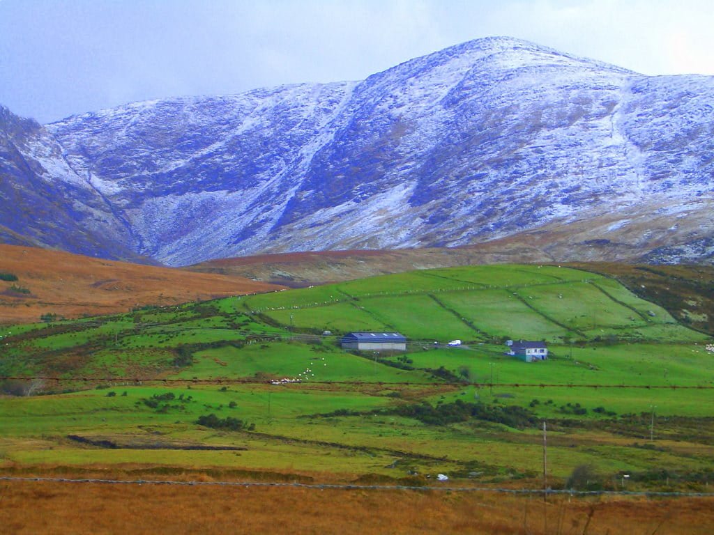 Snow-dusted mountains behind green, patchwork farmland with buildings and scattered sheep.