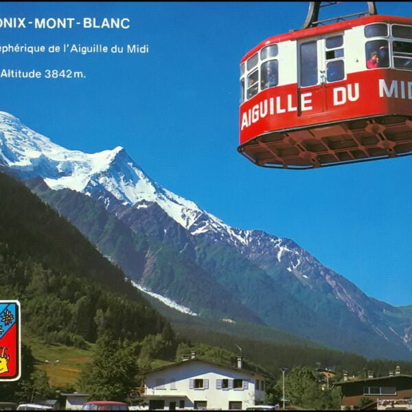 A red cable car with "AIGUILLE DU MIDI" written on the side is suspended in the air against a clear sky, with the snowy peak of Mont Blanc in the background and alpine scenery below, including a village and forested valleys. Text at the top reads "CHAMONIX - MONT - BLANC" and "Départ du téléphérique de l’Aiguille du Midi Altitude 3842m." A coat of arms is in the lower left corner.