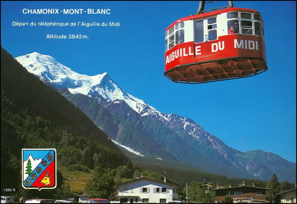 A red cable car with "AIGUILLE DU MIDI" written on the side is suspended in the air against a clear sky, with the snowy peak of Mont Blanc in the background and alpine scenery below, including a village and forested valleys. Text at the top reads "CHAMONIX - MONT - BLANC" and "Départ du téléphérique de l’Aiguille du Midi Altitude 3842m." A coat of arms is in the lower left corner.