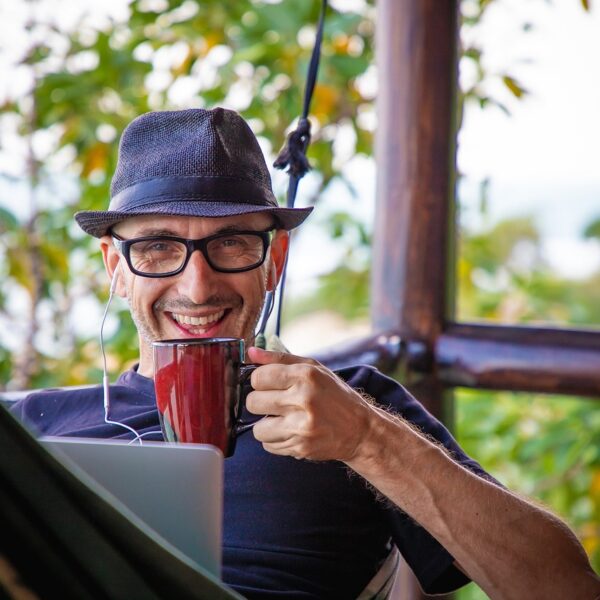 A smiling man wearing a fedora hat and glasses, holding a red mug and using earphones while working on a laptop in a tropical outdoor setting.