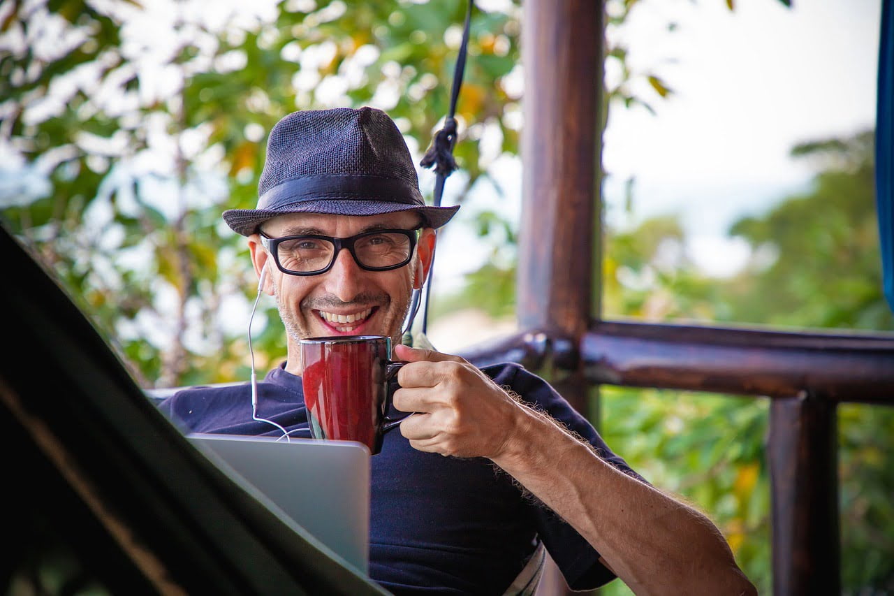 A smiling man wearing a fedora hat and glasses, holding a red mug and using earphones while working on a laptop in a tropical outdoor setting.
