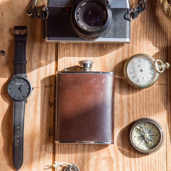 A collection of vintage items laid out on a wooden surface, including a wristwatch, a classic camera, a leather-wrapped flask, pocket watch, compass, and a knife with a decorative handle.