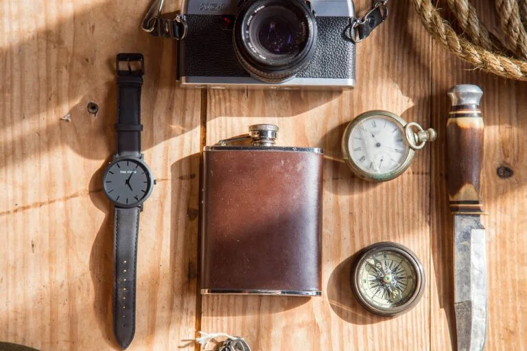 A collection of vintage items laid out on a wooden surface, including a wristwatch, a classic camera, a leather-wrapped flask, pocket watch, compass, and a knife with a decorative handle.