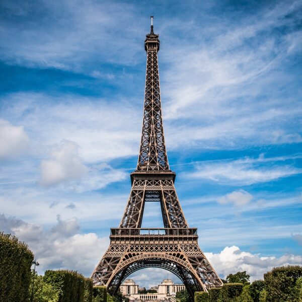 The Eiffel Tower stands prominently against a blue sky with scattered clouds, flanked by green treetops at its base.