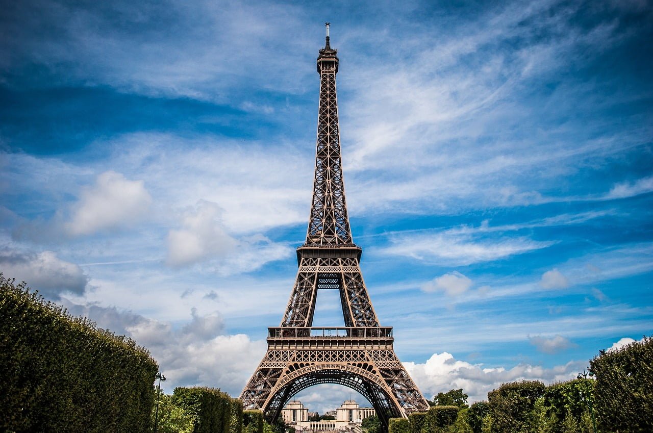 The Eiffel Tower stands prominently against a blue sky with scattered clouds, flanked by green treetops at its base.