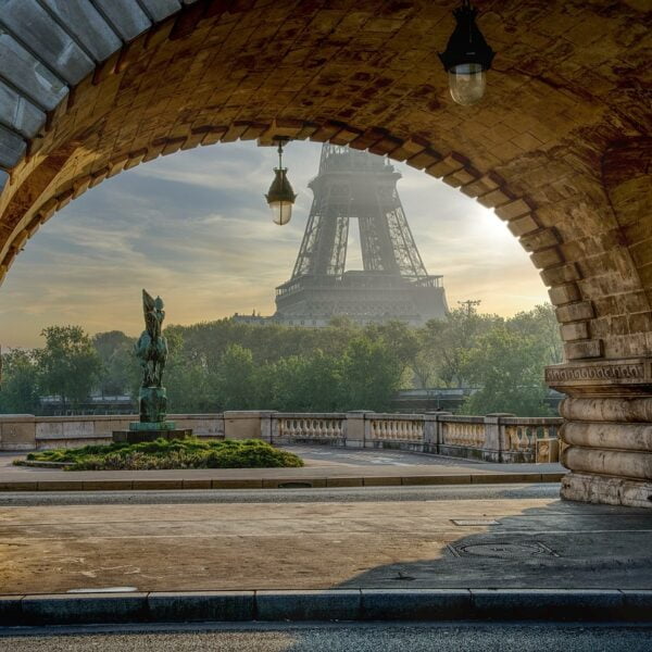 A view of the Eiffel Tower in Paris seen from under an arched stone bridge with ornate street lamps and a bronze statue in the foreground during sunrise.