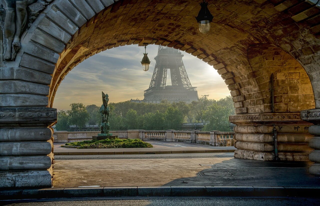 A view of the Eiffel Tower in Paris seen from under an arched stone bridge with ornate street lamps and a bronze statue in the foreground during sunrise.