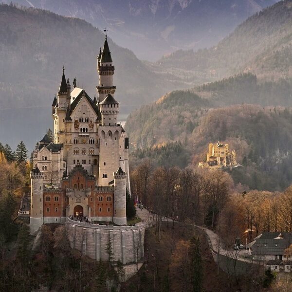 A majestic castle with towering spires and intricate architecture situated on a forested hilltop overlooking a valley with a backdrop of mountain ranges during autumn, with a smaller castle visible in the distance.