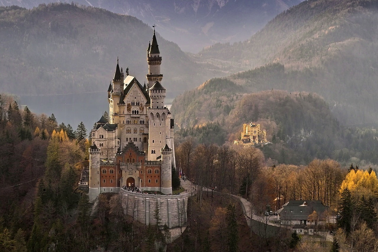 A majestic castle with towering spires and intricate architecture situated on a forested hilltop overlooking a valley with a backdrop of mountain ranges during autumn, with a smaller castle visible in the distance.