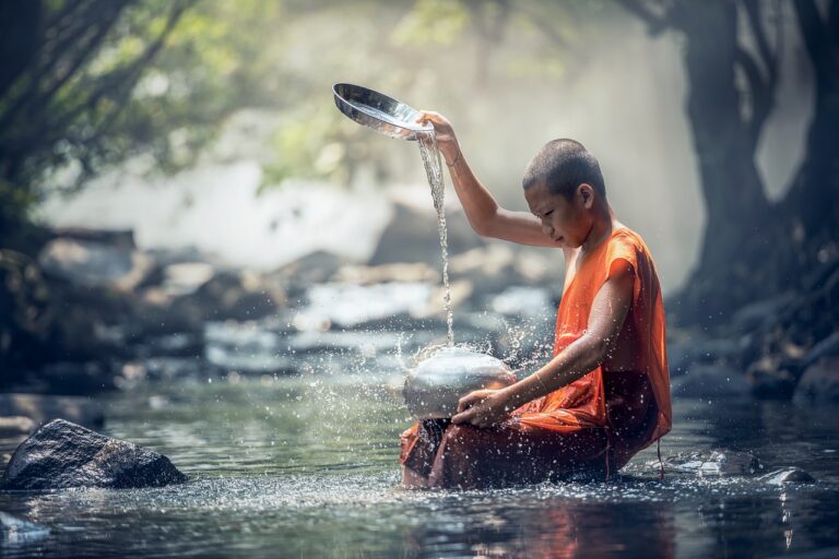 A young monk in an orange robe is sitting on a rock, pouring water from a bowl into a stream surrounded by nature.