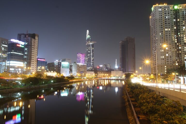A nighttime cityscape showing illuminated skyscrapers reflecting in a calm river, with a clear sky and street lights along a tree-lined promenade.